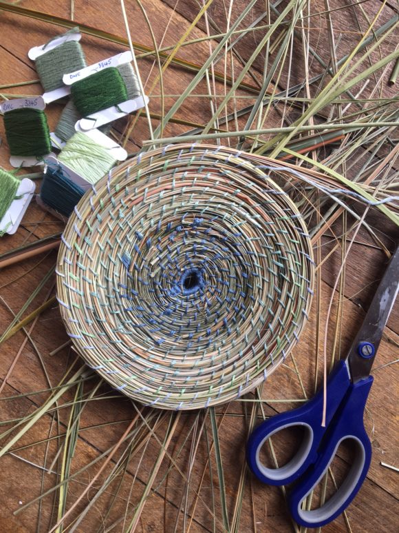 A coiled grass basket in progress with fibres and cordage beside it